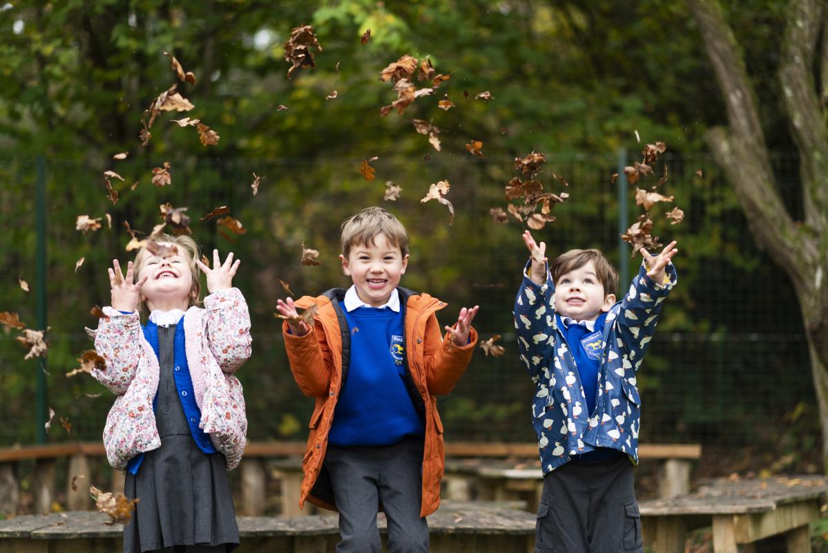 Three Stedham pupils throwing autumn leaves into the air in the woodland beside the school